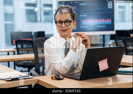 Businesswoman with short gray hair and glasses is seated at a desk in a contemporary office, actively engaging with her laptop, reflecting a professio Stock Photo