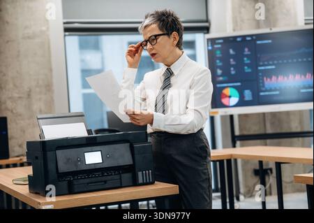 Professional woman in a white shirt and tie is reviewing printed documents at a contemporary office workspace, featuring a printer and a digital displ Stock Photo