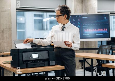Professional woman in a white shirt and tie is operating a printer in a contemporary office, holding printed documents while concentrating on her work Stock Photo