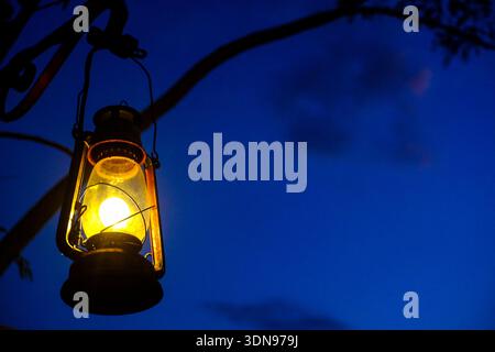 A vintage oil lantern, illuminated and glowing warmly, hangs from a dark tree branch against a deep blue twilight sky. Stock Photo
