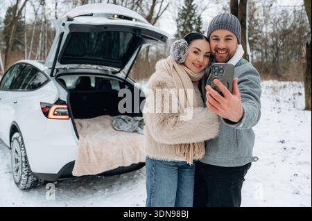 Smiling couple, woman with long dark hair and man with beard, taking selfie in snowy environment beside their car, trunk open revealing cozy blanket, Stock Photo
