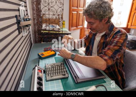 Man in home office repairing a laptop, carefully using a screwdriver on the hardware. Technician fixing modern technology Stock Photo