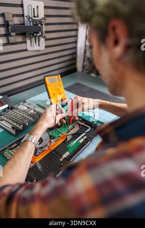Technician performing diagnostics on a laptop device, checking electrical current with a multimeter tool on the circuit board in a workshop Stock Photo