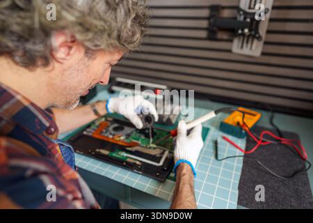 Man wearing gloves soldering components on a laptop motherboard for repair. Focusing on technical work and problem solving Stock Photo