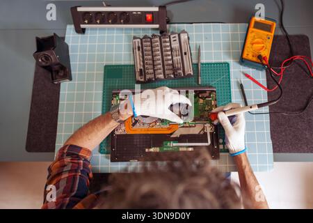 Electronics technician working on a laptop motherboard, performing precise repair with a soldering iron and various technical tools Stock Photo