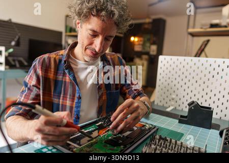 Male technician expertly soldering electronic components on a detailed circuit board, performing specialized computer repair work Stock Photo