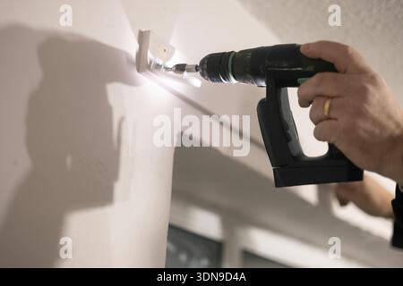 A person’s hand using a cordless power drill to install a roller blind above a window in an interior space. Home renovation. Stock Photo