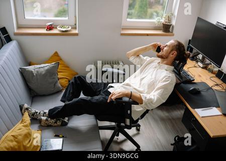 A man is sitting on a sofa in his office, talking on a phone. He has his feet up and a relaxed posture. A work desk with a computer is in the backgrou Stock Photo