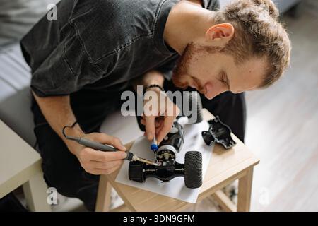 A man is fixing a remote control car model at home during the afternoon. He uses tools and examines the car closely on a table with various parts arou Stock Photo