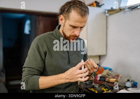A man is focused on a small flashlight in his hands while he works in a workshop. Tools and materials are visible in the background during daytime. Stock Photo