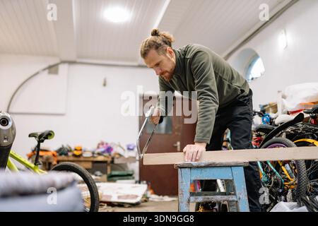 Man focuses on cutting a piece of wood in a workshop filled with bicycles and various tools. The scene shows a busy environment with ongoing projects. Stock Photo