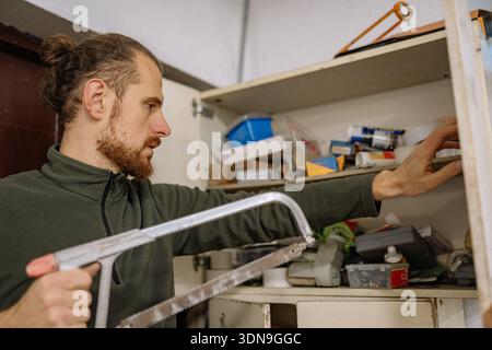 A man sorts through items in a cluttered workshop while holding a saw. He looks focused as he finds what he needs among various tools and materials. Stock Photo