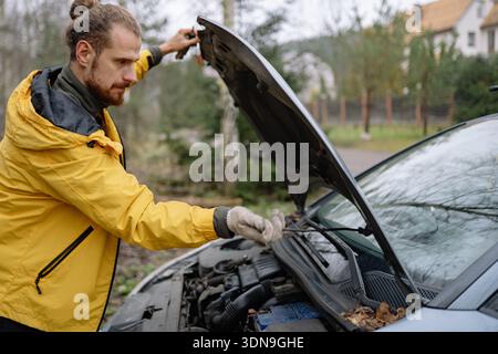A man wearing a yellow jacket repairs a car engine in a rural setting. He stands next to the vehicle with the hood open, focused on the task at hand. Stock Photo