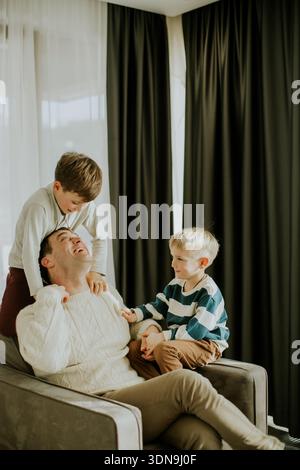 A father sits on a couch while two boys play around him. One boy is climbing on his back, and the other sits next to him. Sunlight comes through the w Stock Photo