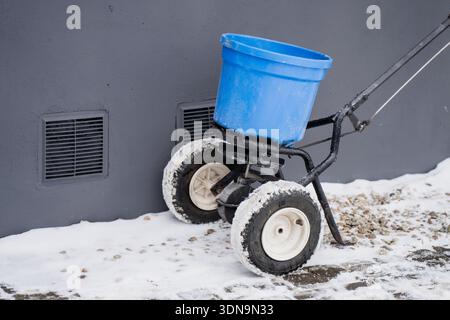Outdoor maintenance equipment, two wheel hand spreader with blue bucket and metal handle, positioned on icy walkway edge beside ventilation grilles an Stock Photo