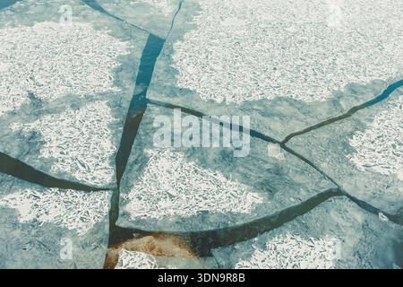 A frozen lake has various cracks and shapes in the ice. Some areas are clear while others hold snow. The patterns show the movement of water below the Stock Photo