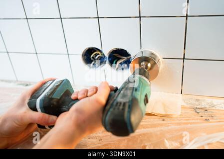 Working person drilling a circular hole into newly installed white ceramic tiles with an electric drill and cutter tool, preparing for electrical outlet installation in a modern kitchen Stock Photo