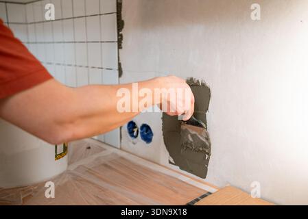 Worker's hand applying adhesive mortar on a white wall using a trowel, installing ceramic tiles in a kitchen or bathroom as part of a home renovation and construction project Stock Photo