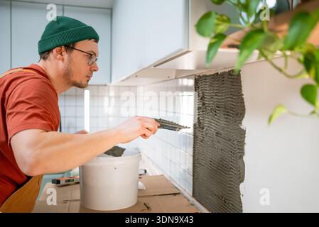 Tiler applying tile adhesive onto a kitchen wall, preparing for ceramic backsplash installation and home renovation, focusing on the details of home interior reconstruction Stock Photo