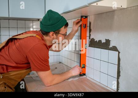 Tradesman installing white ceramic tiles, carefully checking the level of the new backsplash in a modern kitchen, focusing on quality and meticulous workmanship during construction Stock Photo