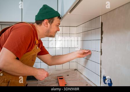 Professional tiler carefully installing new white ceramic tiles on a kitchen wall, demonstrating precision and expertise during home improvement and renovation project Stock Photo