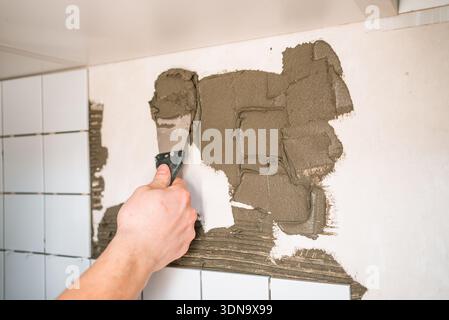 Worker's hand using a trowel to evenly spread cement-based tile adhesive on a kitchen wall, preparing for the precise application of new ceramic tiles during a renovation Stock Photo