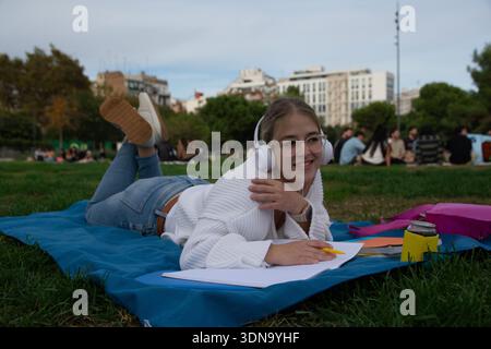 Young woman student lying on a mat in the park, wearing headphones, studying, and smiling, with other students relaxing in the background, representin Stock Photo