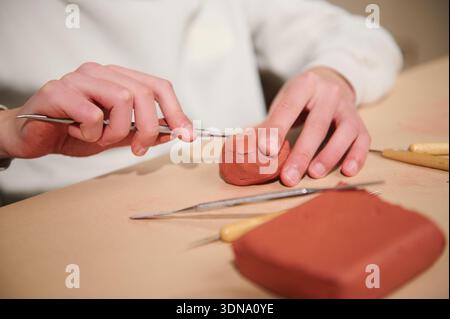 A close view of hands shaping red clay with sculpting tools on a worktable, capturing creativity and focus in a pottery hobby setting. Stock Photo