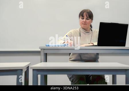 Woman with brown hair is seated at a desk, writing notes and using a laptop in a bright classroom, emphasizing concentration and educational atmospher Stock Photo