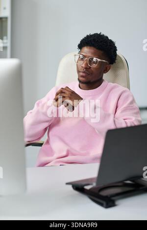 Young Black Man Sitting at Desk Using Computer and Looking Thoughtful Stock Photo