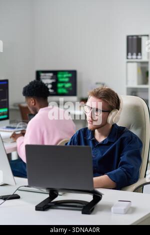 Young Man Wearing Headphones Working on Laptop in Office Setting Stock Photo