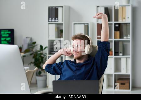 Young Man Wearing Headphones Stretching While Working at Desk Stock Photo