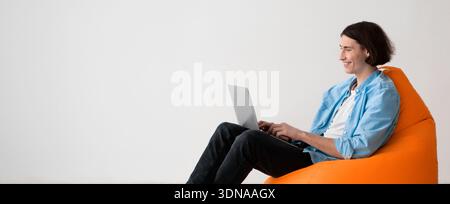 Young Man Works on Laptop While Sitting on Orange Bean Bag Chair in Bright Room Stock Photo