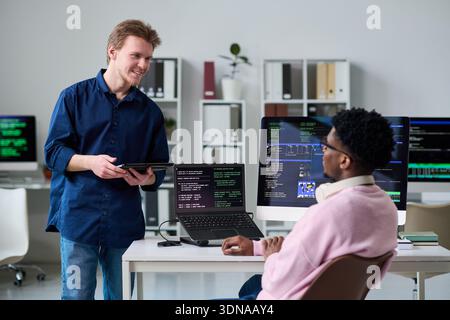 Young Man Talking to Young Black Man Working with Computer Screens Stock Photo