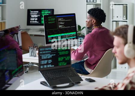 Young Black Man Reviewing Programming Code on Multiple Computer Screens Stock Photo