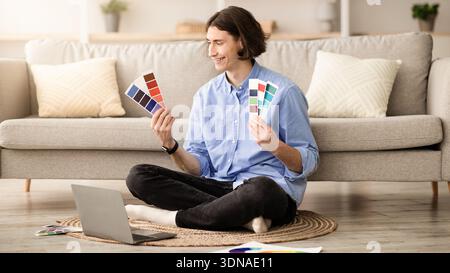 Young Man Sitting on Floor Examines Color Swatches in Front of Laptop in Living Room Stock Photo