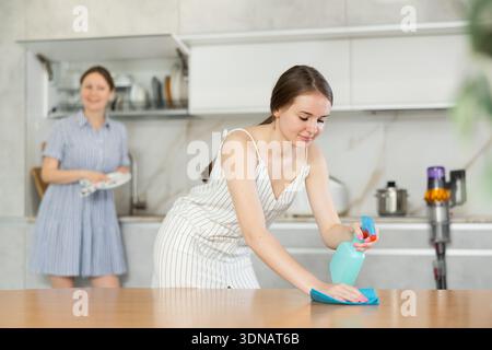 Daughter dusting table while mother washes dishes in kitchen Stock Photo