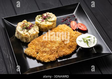 Crispy breaded schnitzel with mashed potatoes and creamy sauce on black plate, dark table Stock Photo
