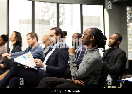 Black young female listening to presentation speech with diverse people in audience, business conference event with speaker. Chairs fill auditorium as participants attend professional summit. Stock Photo