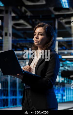 Data center female engineer using laptop, managing server virtualization. Computer scientist using notebook in server room, doing upgrades sustaining long term infrastructure reliability Stock Photo
