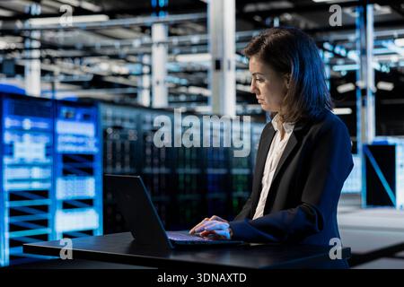 Inspecting infrastructure, laptop and female programmer in data center updating network systems. IT professional using notebook to ensure stable server hub operations, overseeing rigs Stock Photo
