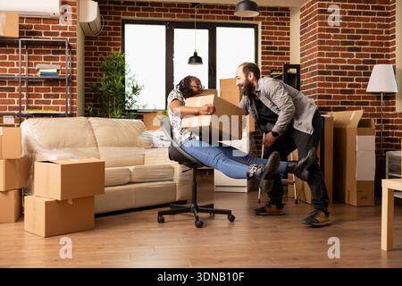 Excited diverse couple celebrating moving day with laughter, as bearded man playfully pushes woman on chair through living room. Cheerful partners enjoying carefree fun in unfurnished apartment. Stock Photo