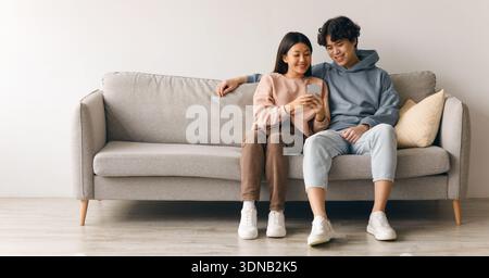 Young Couple Sits Together on Gray Couch Enjoying Time With Smartphone in Cozy Indoor Space Stock Photo