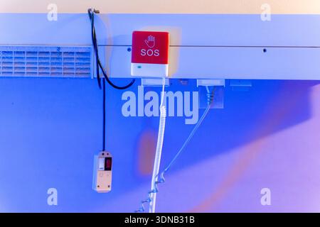 Red SOS emergency call button with hand icon and nurse call remote mounted on blue illuminated wall in hospital room. Concept of patient safety, medic Stock Photo