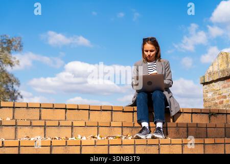 Focused young female freelancer working on laptop while sitting on urban steps. Stock Photo