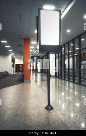 Vertical blank lightbox billboard mockup on pole in modern airport terminal corridor with glossy floor, ceiling lights and glass wall, empty illuminat Stock Photo
