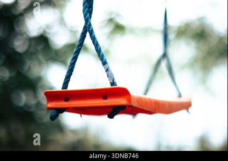 Close-up of an empty orange plastic swing seat hanging from blue ropes in a garden, featuring a soft bokeh background of trees. Stock Photo