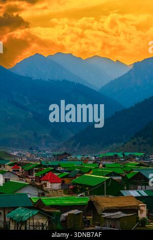 Green rooftops of houses in Sonamarg / Sonmarg, nestled between towering Himalayan mountains. Orange rays of setting sun lights up the monsoon clouds. Stock Photo