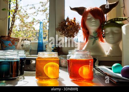 Sunlit kitchen craft scene with easter eggs soaking in a glass jars of yellow and orange food dye near a window Stock Photo