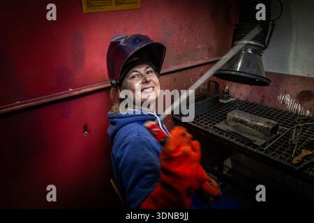 Smiling female welder holding a welding torch in an industrial workshop, protective helmet and gloves, metalworking environment. Stock Photo